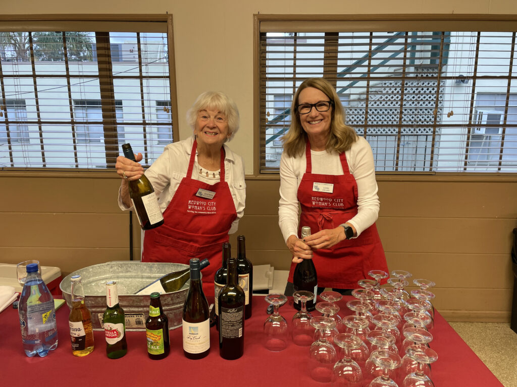 Two women are wearing red aprons and smiling at the cameral. One woman is holding up a bottle of wine, and the other woman is open a bottle of Prosecco