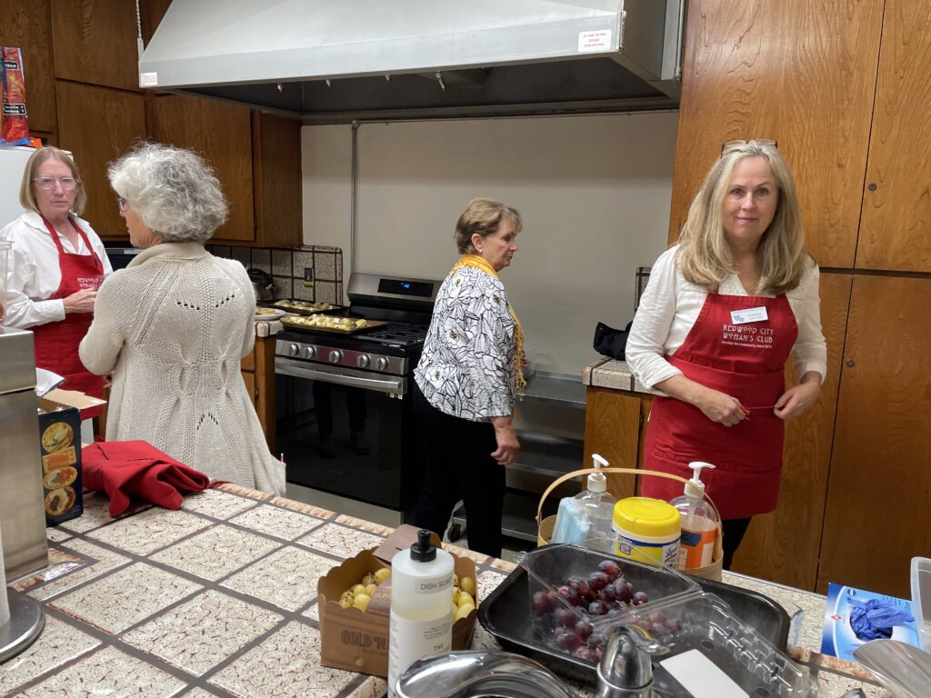 Four women are in a kitchen preparing food. Two women are wearing red aprons with Redwood City Woman's Club in white embroidery.