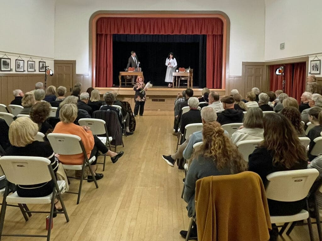 Two people are on a stage. A woman stands in front of them with a mic. An audience is watching them.