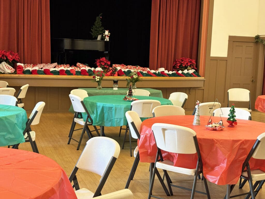 Auditorium filled with round tables covered in green and red tablecloths. A stage is decorated with Christmas decorations.