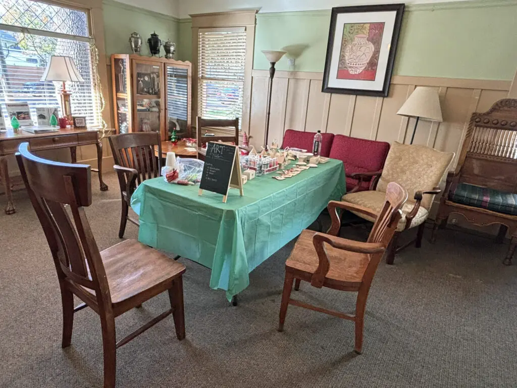 A room has a rectangular table in the center with a green table cloth and art supplies. Wooden chairs are set up around it.