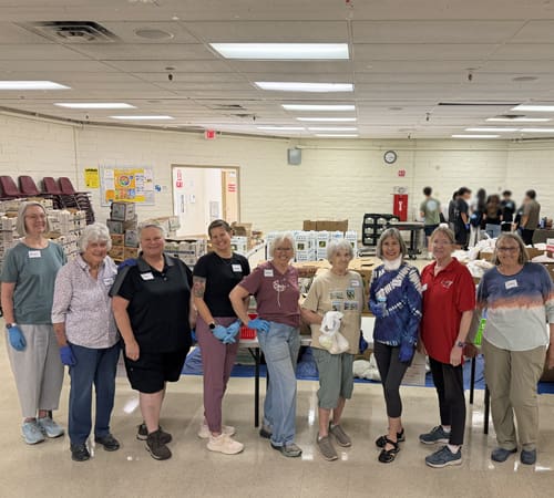 A group of women are facing the camera. Behind them is a room of food for distribution